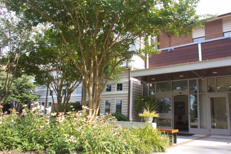 Entrance of a senior living facility with a garden full of flowering plants and trees in front. The building has large windows and a covered entryway with glass doors.