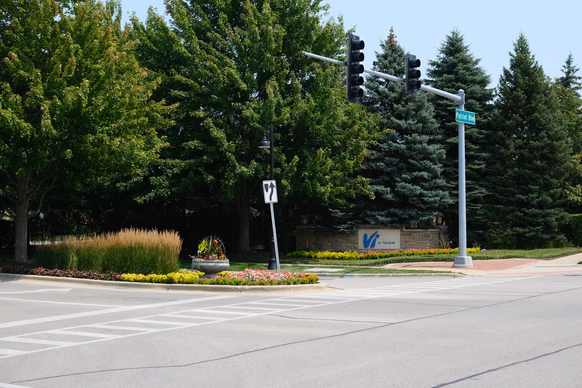 Street intersection with landscaped median and the 'Vi at The Glen' entrance sign, traffic lights, and trees in the background.