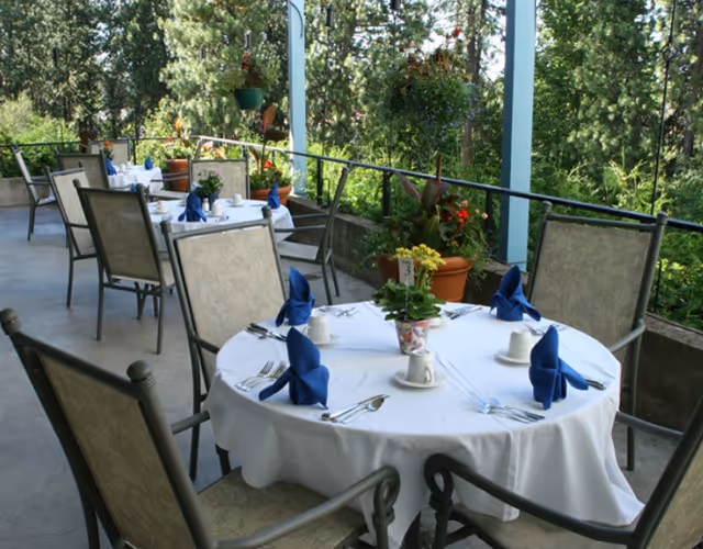 Outdoor dining area with round tables covered in white tablecloths, each set with blue folded napkins, cups, and silverware. The area is surrounded by greenery and potted plants, with a railing along the edge and tall trees in the background.