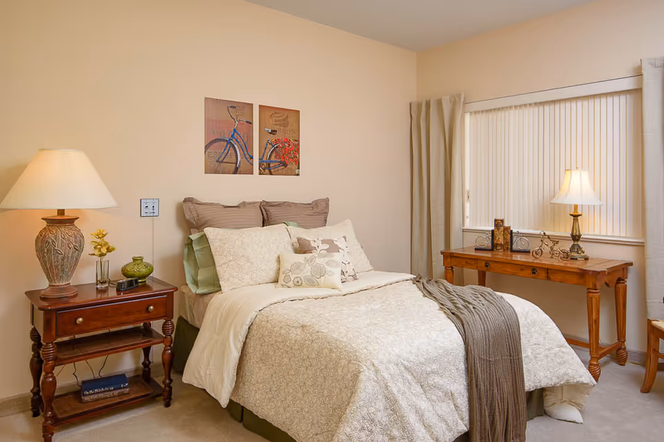 A cozy bedroom with a neatly made bed featuring multiple pillows and a beige patterned comforter. There is a wooden nightstand with a decorative lamp, a small vase with yellow flowers, and a green decorative item. On the wall above the bed are two framed pictures of a bicycle with flowers. To the right, a wooden console table holds a lamp and small decorative bicycle figurines. The window behind the table has vertical blinds and beige curtains.