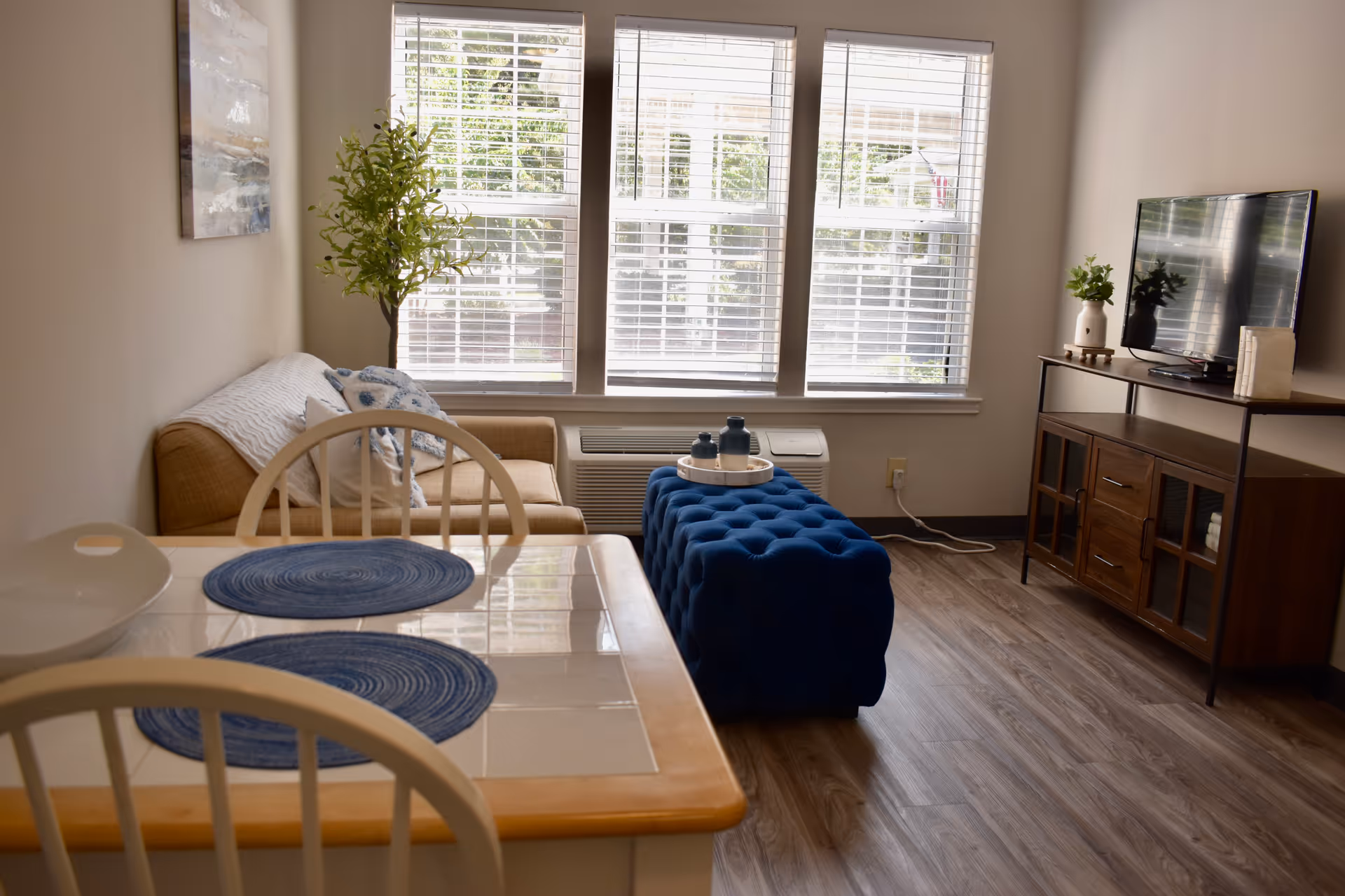 Sunlit living room and dining area with a small table, beige sofa, blue tufted ottoman, TV on a wooden stand, and three large windows with blinds.