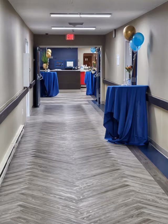 A hallway in Dennett Rehab Center decorated with blue and gold balloons attached to tables draped in blue cloth. The hallway has light-colored walls, a patterned floor, and leads to a reception area with a dark counter and blue wall in the background. An exit sign is visible above the doorway.