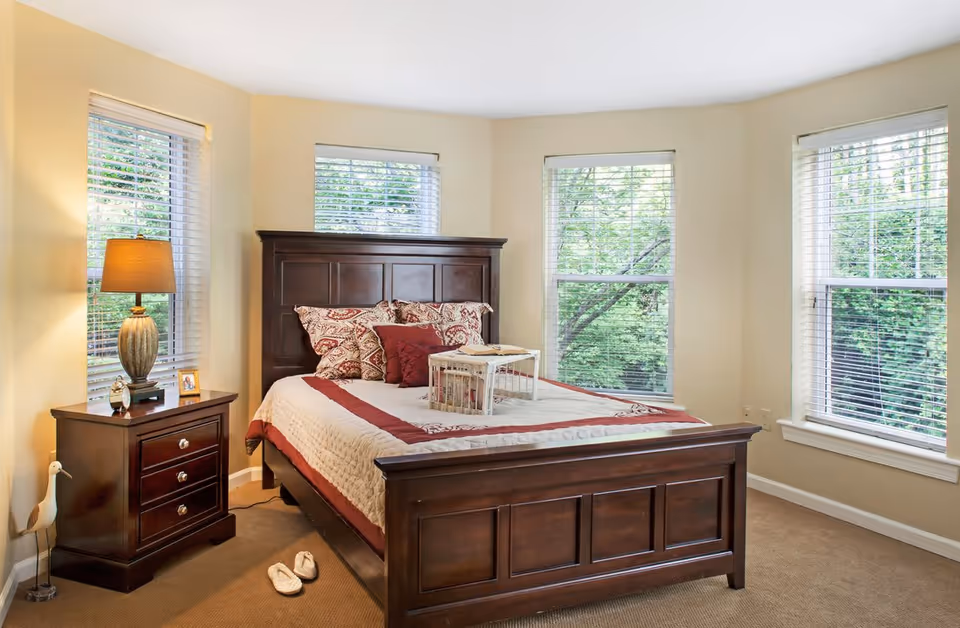 Sunlit bedroom with a dark wooden bed, matching nightstand and lamp, three windows with blinds, and decorative pillows.