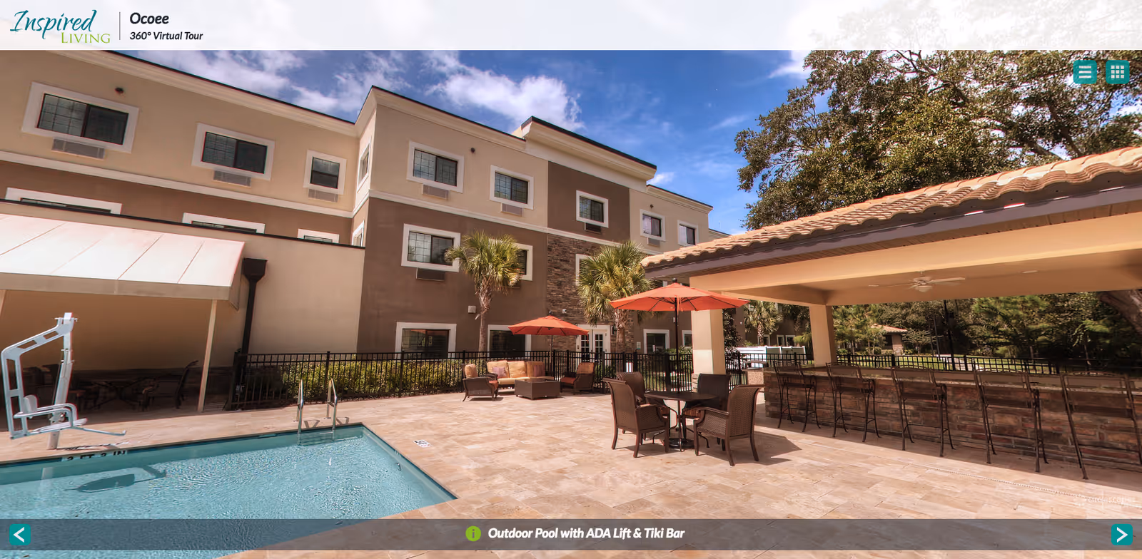 Outdoor area of Inspired Living Ocoee featuring a swimming pool with an ADA lift, patio seating with tables and umbrellas, a tiki bar with bar stools under a covered area, and a multi-story building in the background under a blue sky with some clouds.