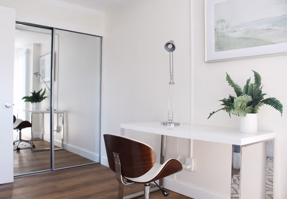 A modern, minimalist room featuring a white desk with chrome legs, a contemporary wooden chair with a white seat, a silver desk lamp, and a potted green plant. There is a large framed artwork on the wall above the desk and a mirrored sliding closet door reflecting part of the room. The floor is wooden and the walls are painted white.