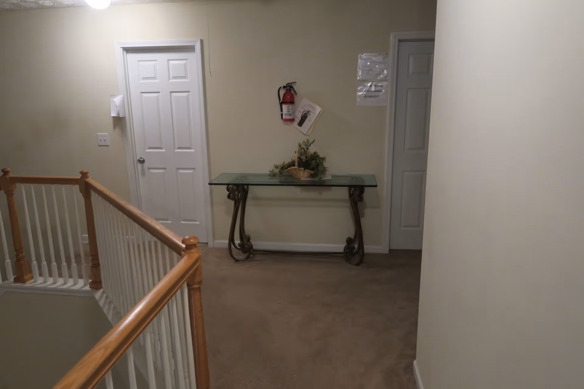Interior hallway with beige walls and carpet, featuring a wooden railing on the left side, two closed white doors on the far wall, a glass-top table with decorative wrought iron legs and a basket with greenery on top, and a fire extinguisher mounted on the wall above the table.