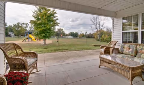 Covered patio area with wicker furniture including a sofa, two chairs, and a glass-top coffee table. The patio overlooks a grassy field with a playground featuring a slide and climbing structure. Trees and houses are visible in the background under a cloudy sky.