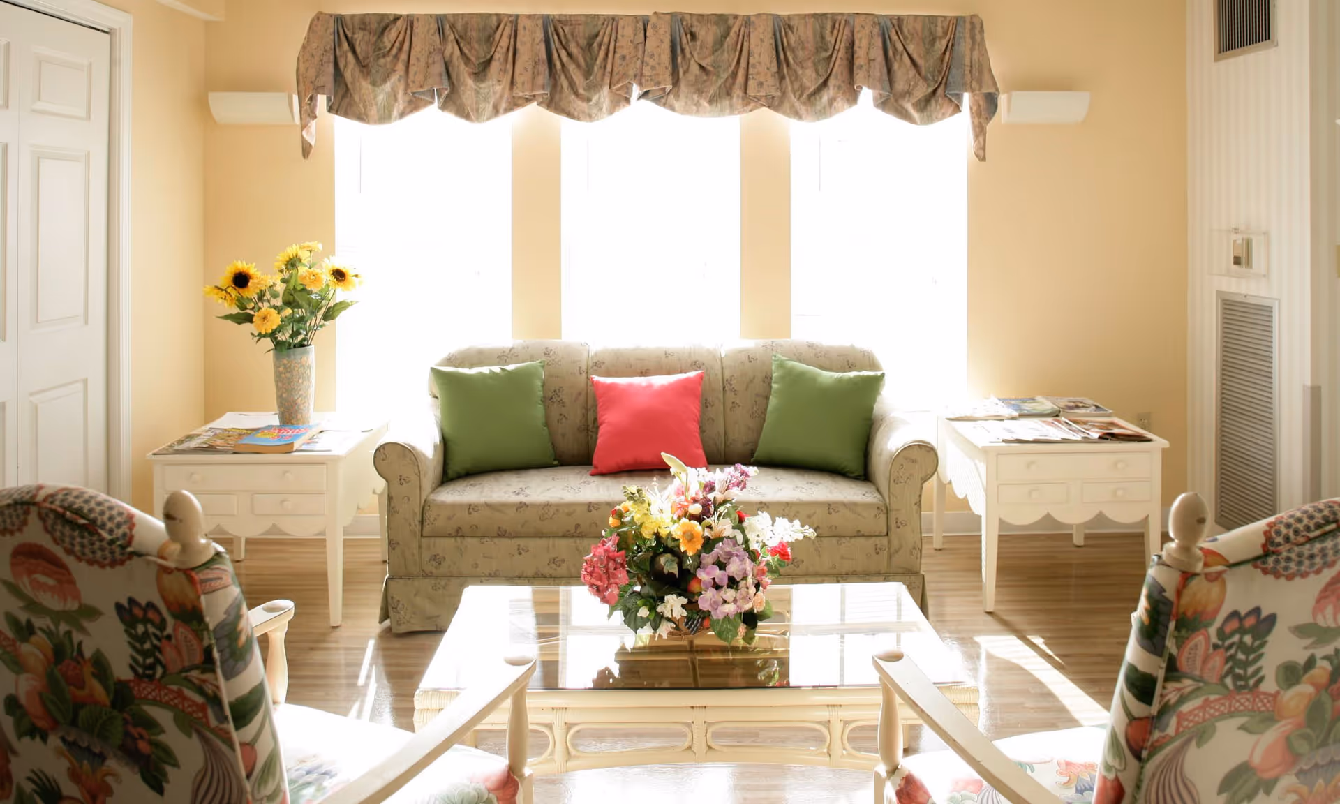 A bright and cozy living room with a floral patterned sofa adorned with two green pillows and one red pillow. In front of the sofa is a glass coffee table with a colorful flower arrangement. Two floral upholstered armchairs face the sofa. On either side of the sofa are white side tables, one with a vase of sunflowers and the other with magazines. Large windows with floral valances allow natural light to fill the room.