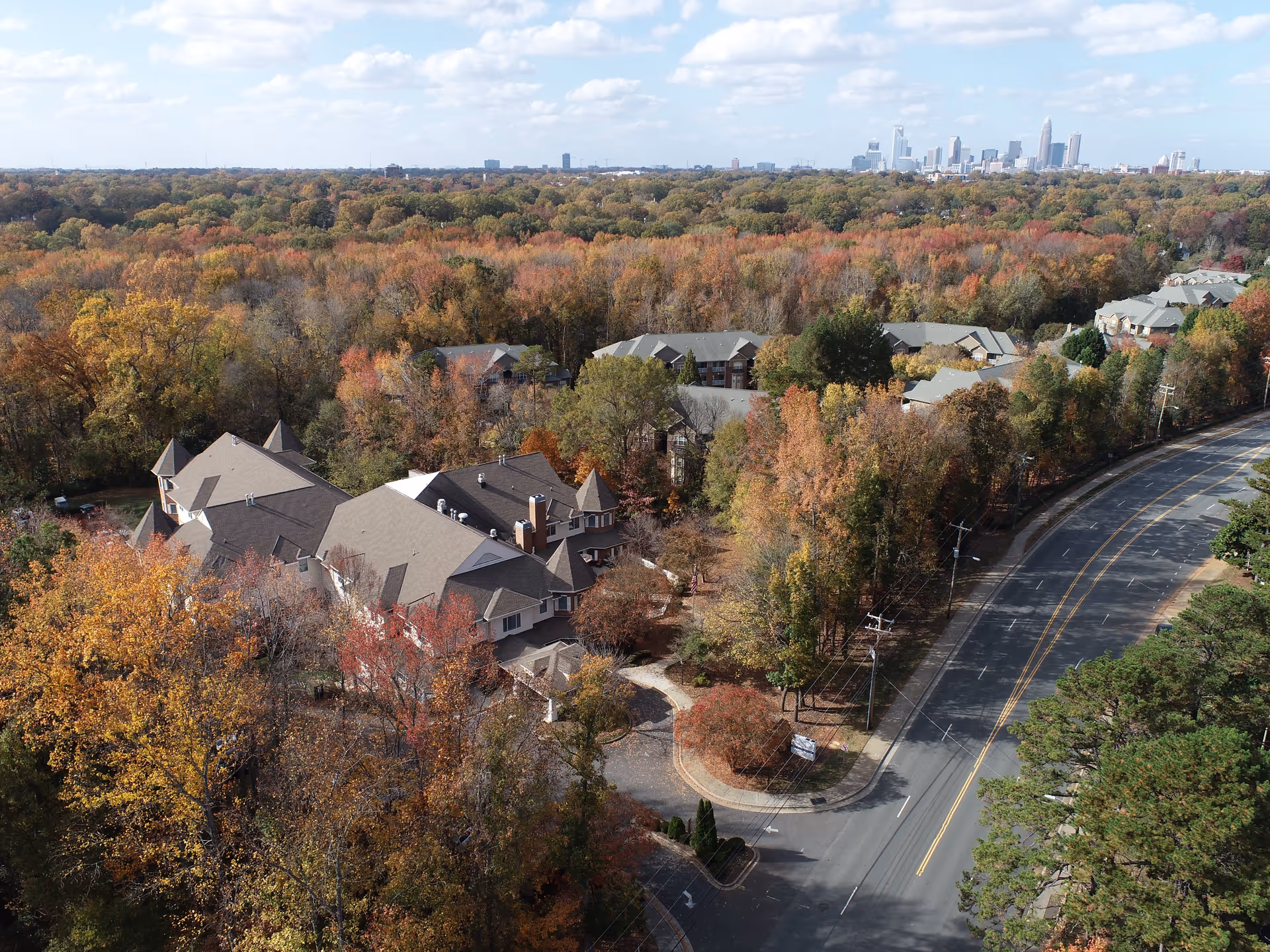 Aerial view of Charter Senior Living of Charlotte surrounded by dense trees with autumn foliage. The facility buildings have pitched roofs and are situated near a curved road. In the background, the skyline of Charlotte city is visible under a partly cloudy sky.