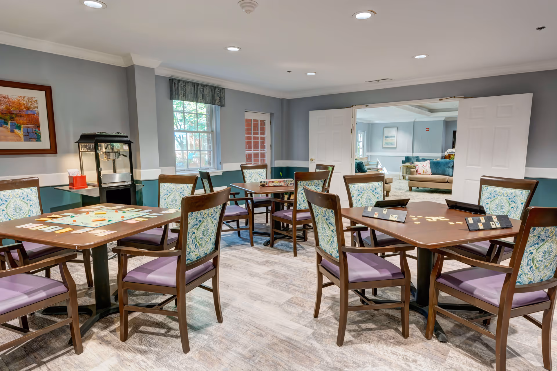 A well-lit activity room with several wooden tables and chairs arranged for playing board games. The chairs have patterned backs and purple cushions. One table has a Monopoly game set up, and another has a Scrabble game in progress. The room has light gray walls with white trim, a window with a valance, a popcorn machine in the corner, and an open doorway leading to a lounge area with sofas and cushions.