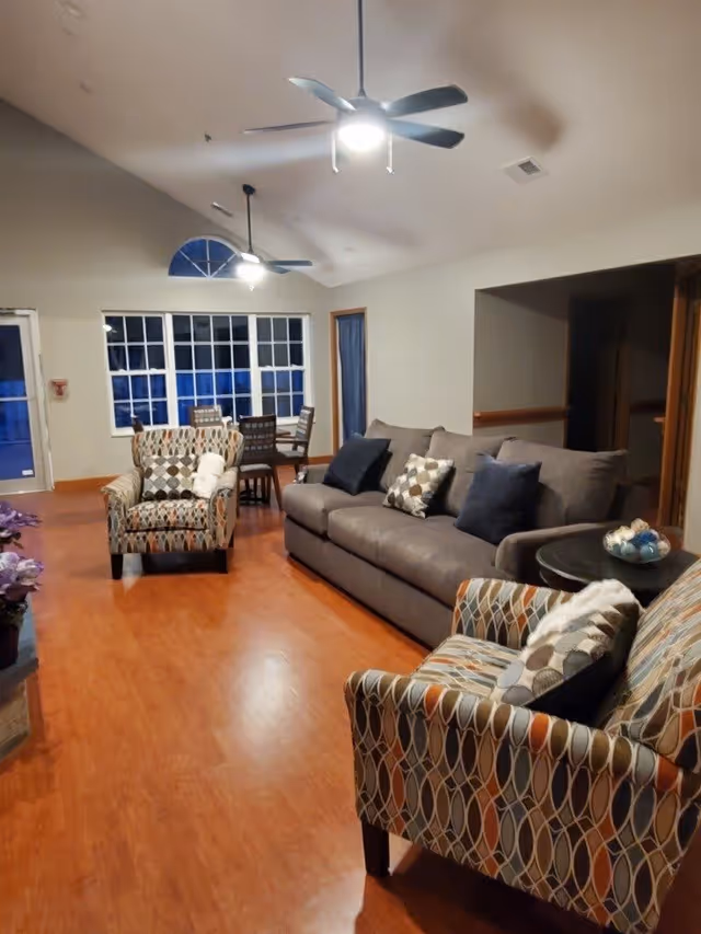 Spacious common living room with a gray sofa, patterned armchairs, a small dining table, ceiling fans, and a large window.