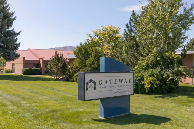 Outdoor view of Gateway Transitional Care Center sign on a grassy lawn with trees and a building with a red roof in the background under a clear blue sky.