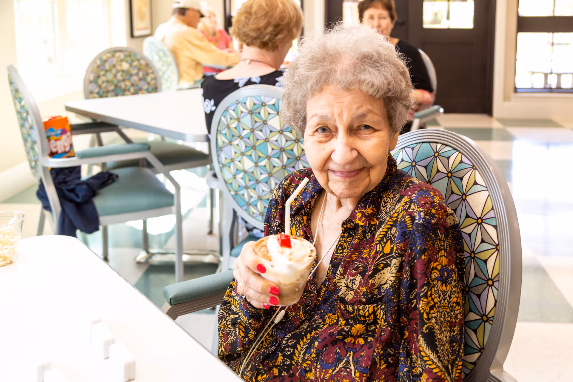 An elderly woman with curly gray hair and a colorful patterned blouse is sitting in a chair with a geometric patterned backrest, smiling and holding a dessert topped with whipped cream and a cherry. In the background, other elderly people are seated at tables in a bright room with large windows.