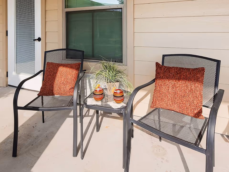 Two black metal mesh chairs with rust-colored cushions on a concrete patio next to a small black metal mesh table holding a potted plant and two striped decorative jars. The patio is adjacent to a beige building with a window and a door with blinds.