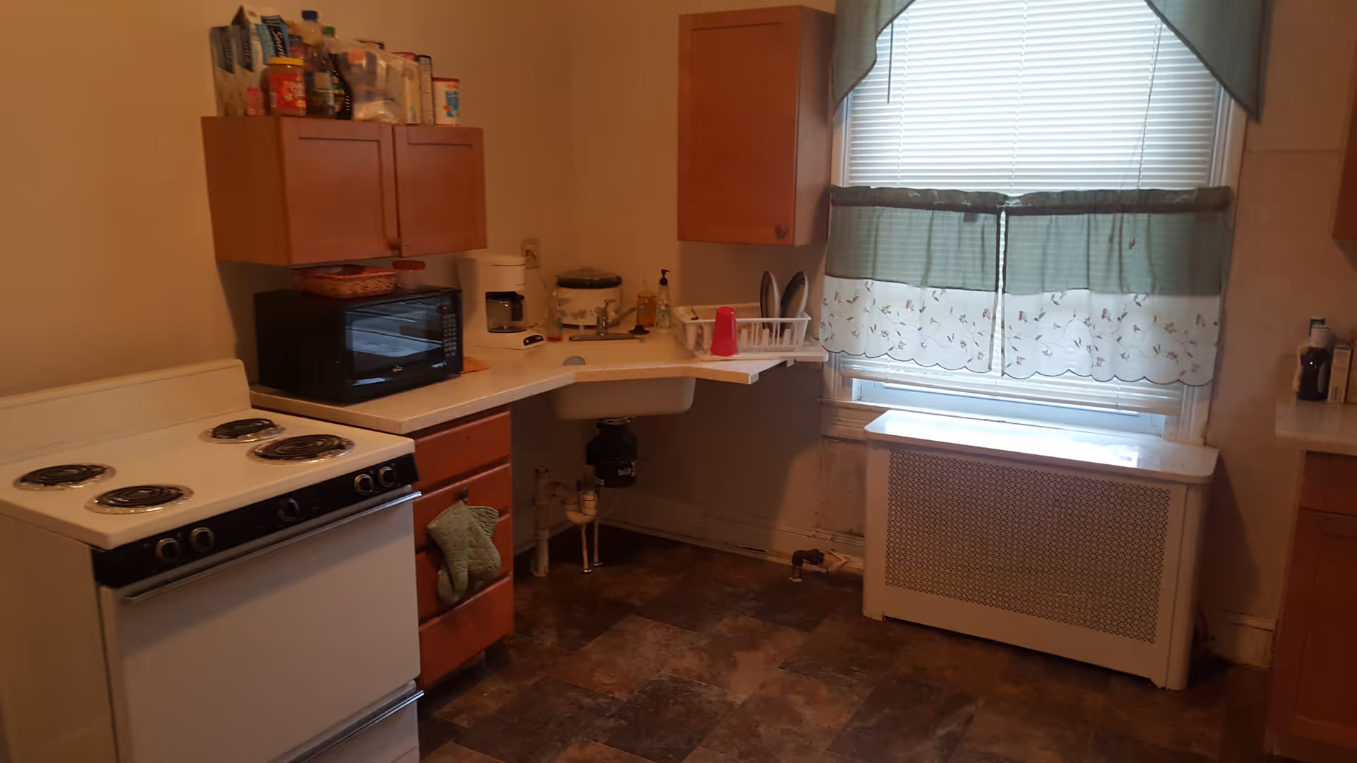 A small kitchen with a white electric stove, wooden cabinets, a microwave, a rice cooker, and a sink. There is a window with green and white curtains above a radiator cover. Various kitchen items are placed on the countertops and cabinets.