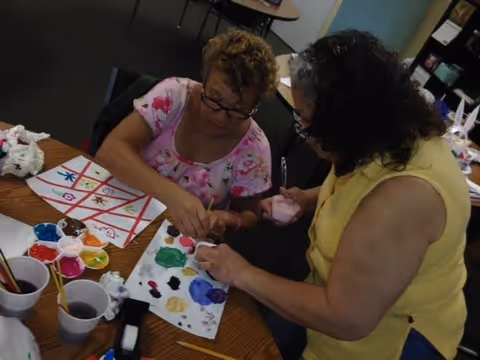 Two women engaged in a painting activity at a table. One woman is wearing a pink floral shirt and glasses, while the other is wearing a yellow sleeveless top. The table is covered with paint palettes, brushes, cups of water, and a paper with colorful painted designs.
