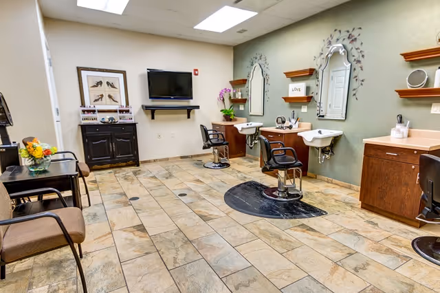 Well-lit salon room with styling chairs, wash basins, mirrors, a wall-mounted TV, and waiting chairs.