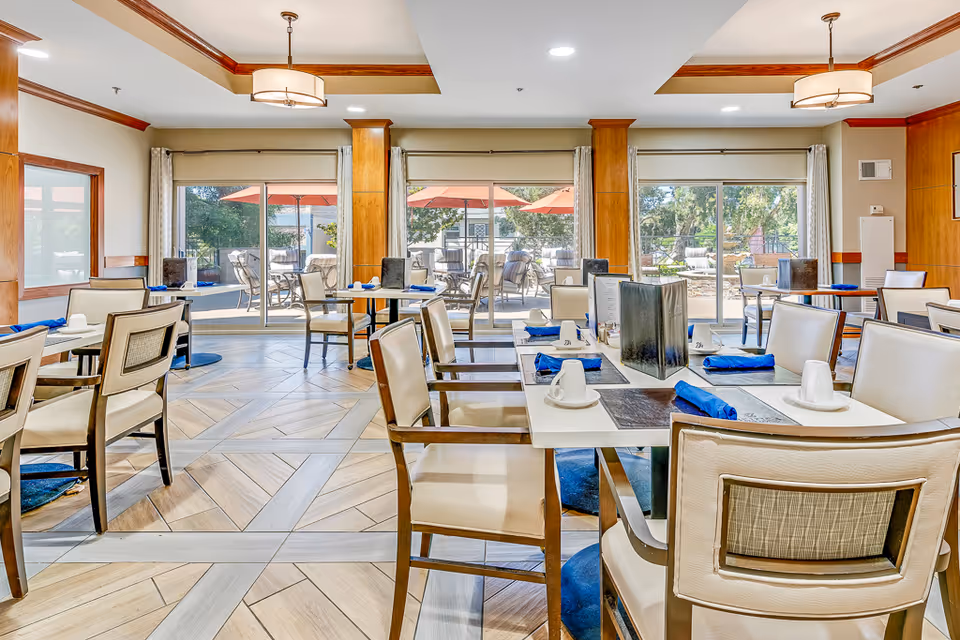 Bright dining room with neatly set tables and chairs, blue napkins and cups, and large sliding glass doors opening to an outdoor patio.
