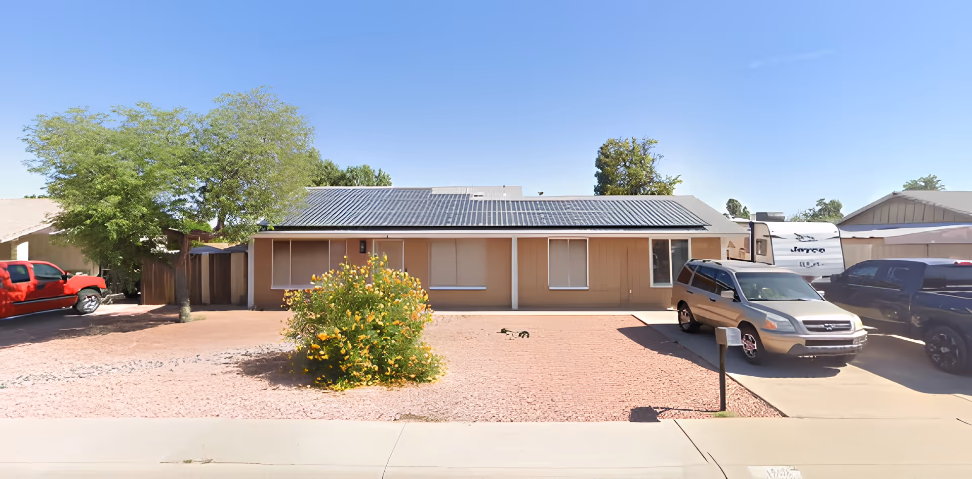 Single-story building with solar panels on the roof, beige exterior walls, and several windows with closed blinds. In front of the building is a gravel yard with a flowering bush in the center and a concrete driveway with parked vehicles, including a silver SUV and a red truck. A tree is visible on the left side of the yard under a clear blue sky.
