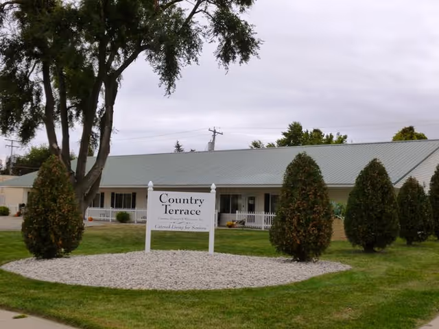 Exterior view of Country Terrace, a single-story building with a gray metal roof, white siding, and a white picket fence. The foreground features a landscaped area with small bushes and a white sign that reads 'Country Terrace, Assisted Living & Memory Care, Central Doors for Seniors.' Trees and a cloudy sky are visible in the background.