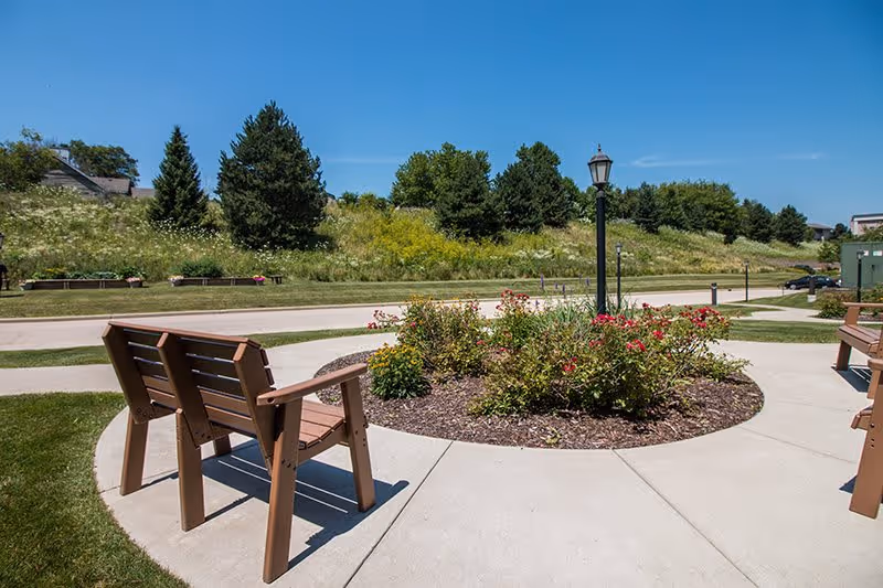 Outdoor seating area with wooden benches arranged around a circular flower bed with blooming plants and a lamp post in the center. The area is surrounded by a paved walkway and grassy landscape with trees and shrubs under a clear blue sky.