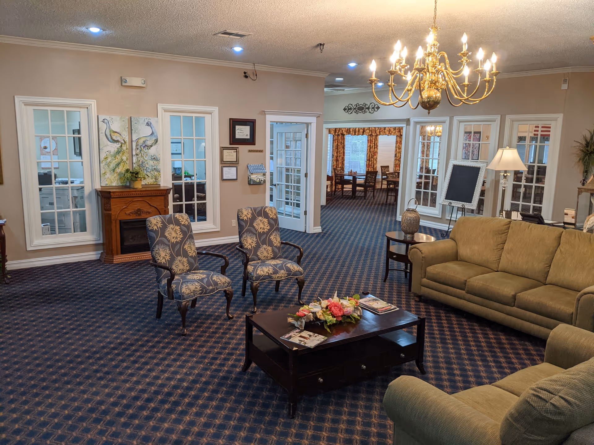 A cozy living room area in a senior living facility with patterned carpet, two upholstered armchairs with floral designs, a wooden coffee table with a flower arrangement and magazines, and two green sofas. The room features a chandelier, a floor lamp, a small electric fireplace with peacock artwork above it, and glass-paneled doors leading to another room with dining tables and chairs.