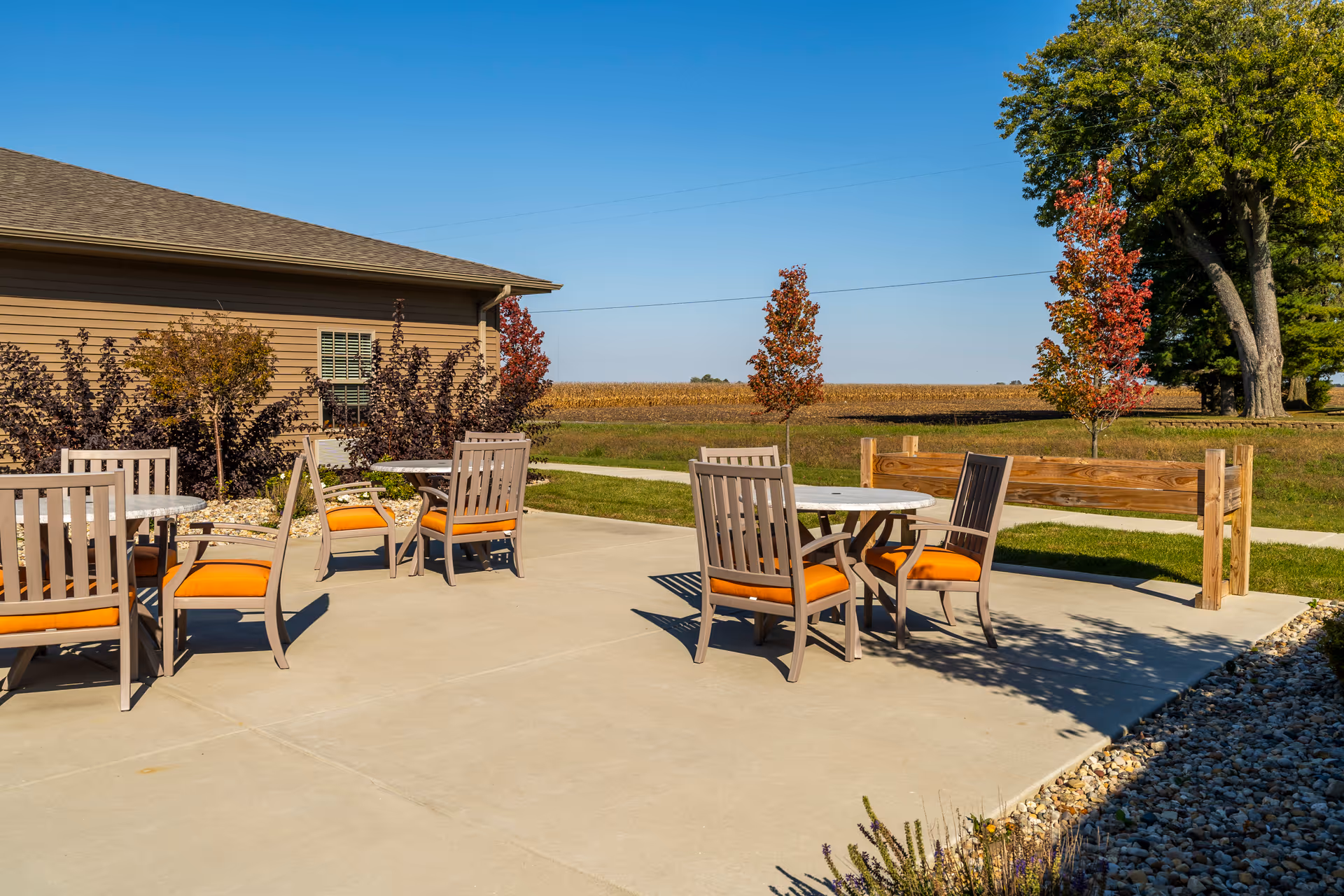 Outdoor patio with round tables and wooden chairs with orange cushions next to a single-story building and open field under a clear blue sky.