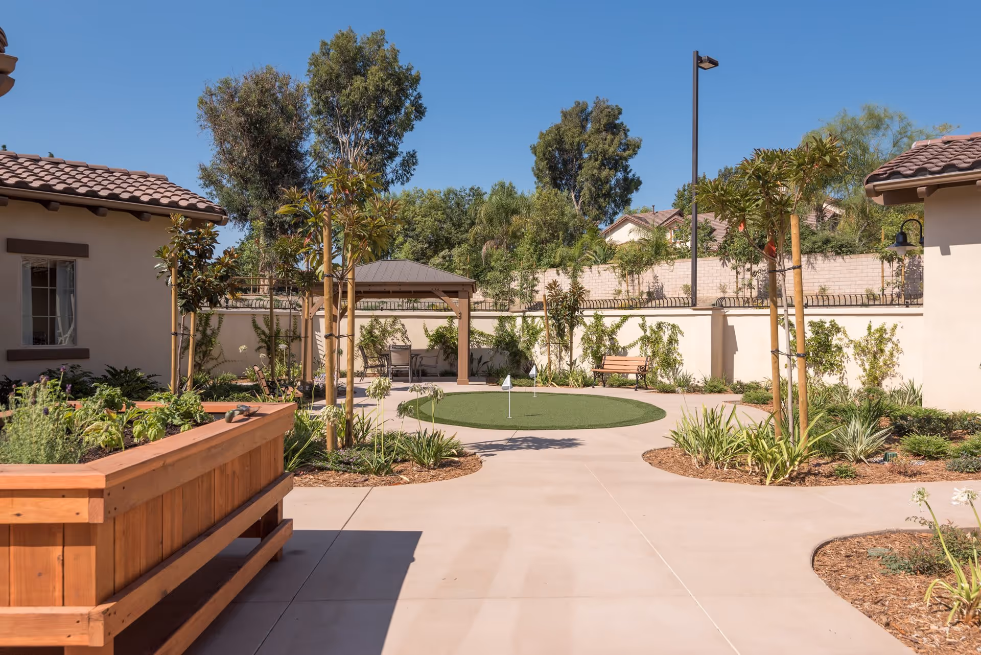 Sunlit courtyard at a senior living facility featuring a small putting green with a flag, gazebo, raised wooden planter, benches, and landscaped paths between single-story buildings.