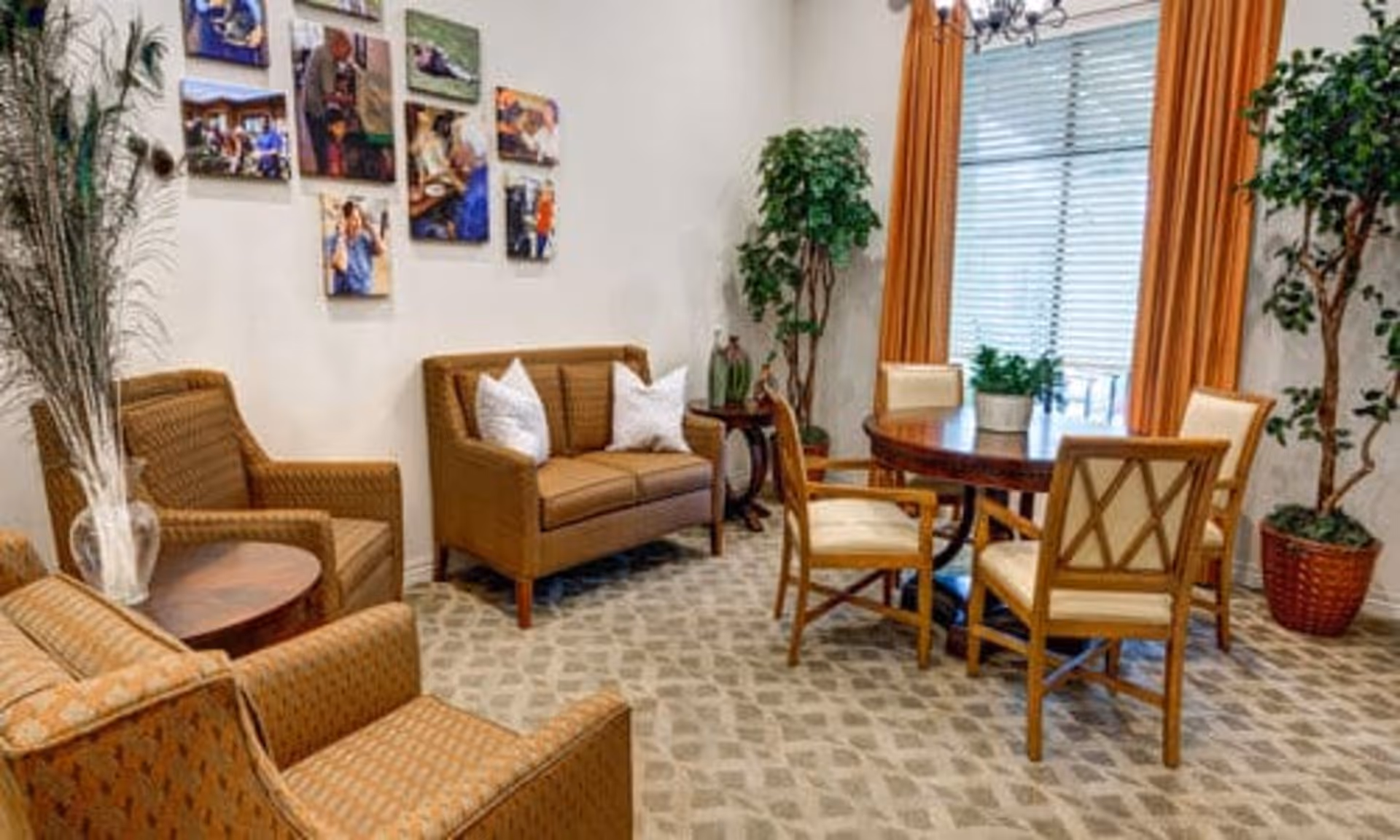 A cozy sitting area with two armchairs and a loveseat with white pillows arranged around a small round wooden table. Nearby is a round dining table with four wooden chairs with cream cushions. The room has patterned carpet, large windows with orange curtains, and several potted plants. A wall displays a collage of framed photos.