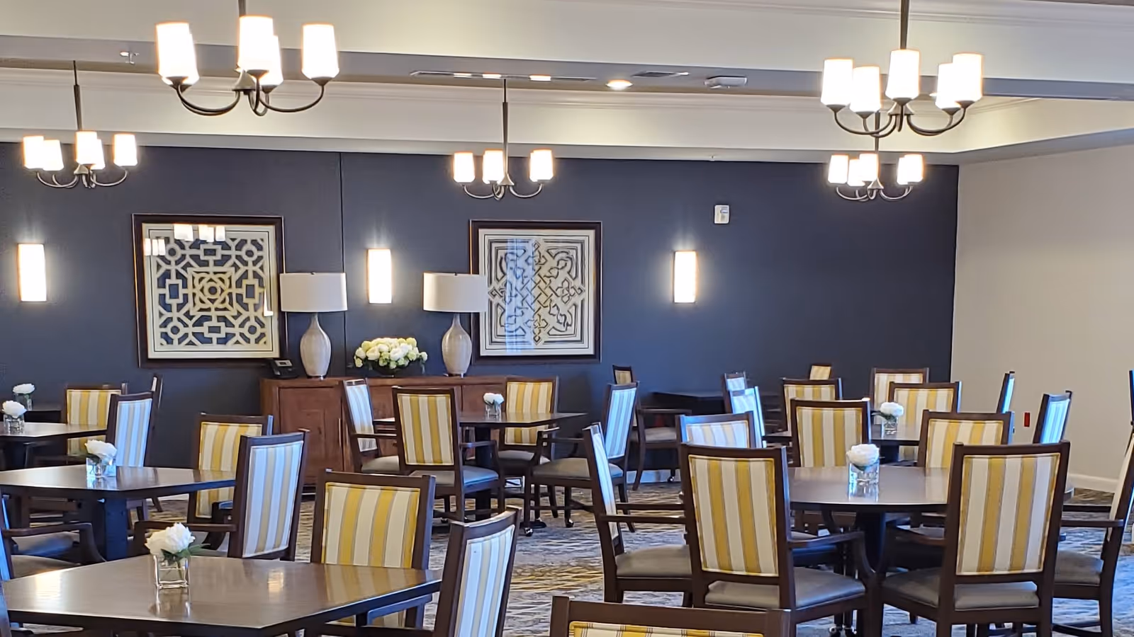 A dining room with multiple round and square tables surrounded by wooden chairs with yellow and white striped upholstery. The room has a dark blue accent wall with two framed geometric artworks, two table lamps, and wall sconces. Several chandeliers hang from the ceiling, providing warm lighting. Small floral arrangements are placed on each table.