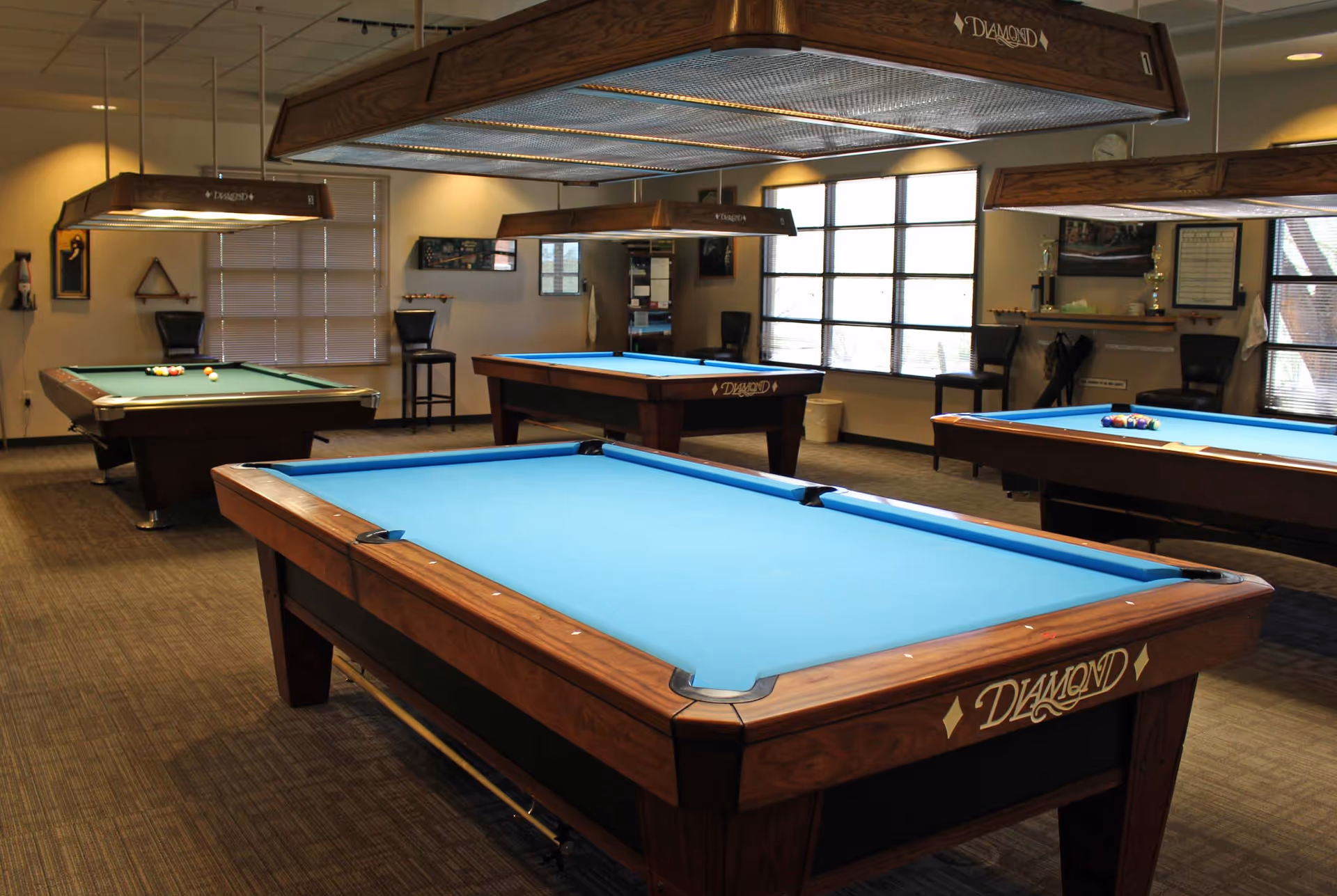 A well-lit indoor billiards room with several pool tables and hanging overhead lights.