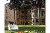 Exterior of a brick multi-story senior living building with a lawn, white bench, flowerbed, and tall trees.