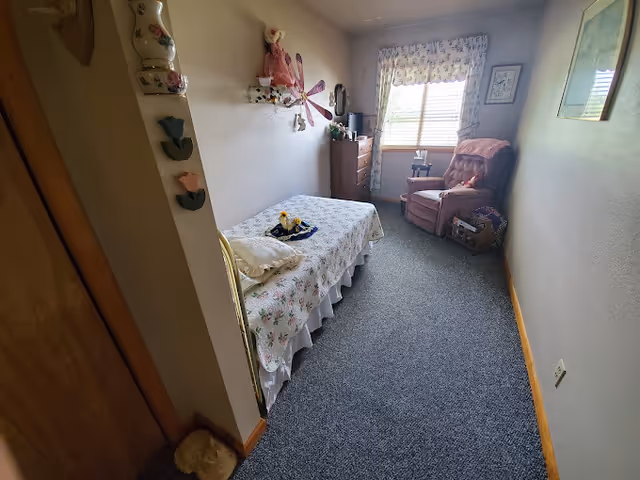 A small bedroom with a single bed covered in a floral bedspread, a pink recliner chair near a window with floral curtains, a wooden dresser, and various decorative items on the walls and shelves.