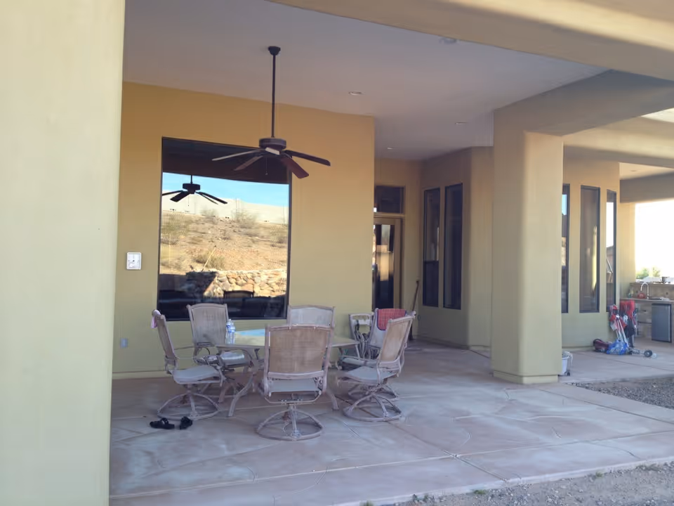 Covered outdoor patio area with a round glass table and six swivel chairs. Ceiling fans hang from the roof, and large windows reflect the outdoor landscape. The patio floor is concrete with a textured pattern, and there is some outdoor equipment and a small grill visible in the background.