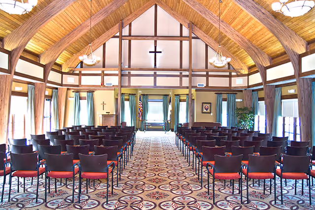 Interior of a chapel or meeting room with rows of black chairs with red cushions arranged facing a wooden podium and a large cross mounted on the wall. The room has a high wooden vaulted ceiling with exposed beams, hanging light fixtures, patterned carpet, and windows with blue curtains.