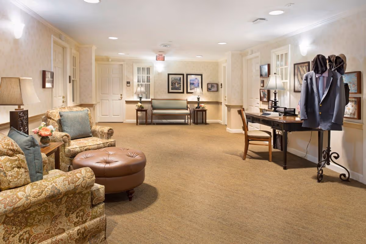 A well-lit interior common area with patterned armchairs, a round leather ottoman, a wooden desk with a chair, and a coat rack holding a suit jacket. The walls are decorated with framed pictures and there are table lamps providing warm lighting. The carpet is beige and the space has multiple doors and windows.
