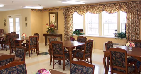 A dining room with multiple wooden tables and chairs featuring floral upholstery. The room has large windows with floral valances, cream-colored walls, and floral centerpieces on the tables. There is a small table with a flower arrangement near the door.