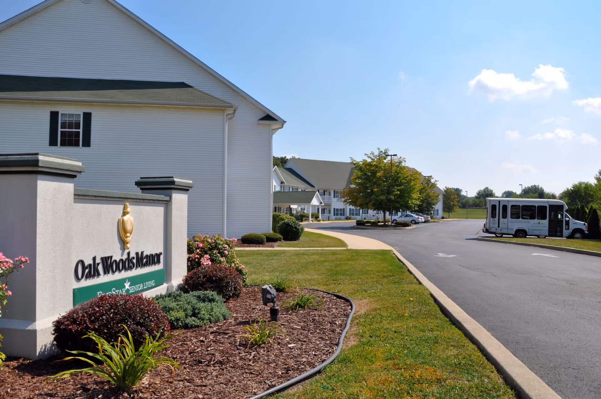 Front entrance of Oak Woods Manor showing the facility sign, landscaped grounds, driveway, and a parked shuttle bus.