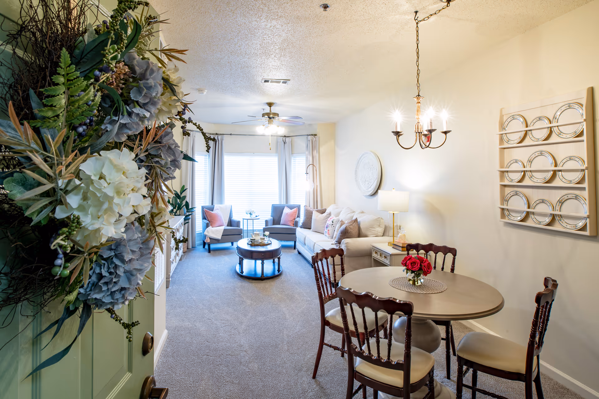 View into a cozy living and dining area of a retirement community apartment. The foreground shows a round dining table with four wooden chairs and a small vase of red roses. The background features a sitting area with a beige sofa, two gray armchairs with pink pillows, a round coffee table, and a large window with white curtains. A ceiling fan with lights and a chandelier above the dining table illuminate the space. A decorative plate rack with plates is mounted on the wall, and a floral wreath is partially visible on the green door.