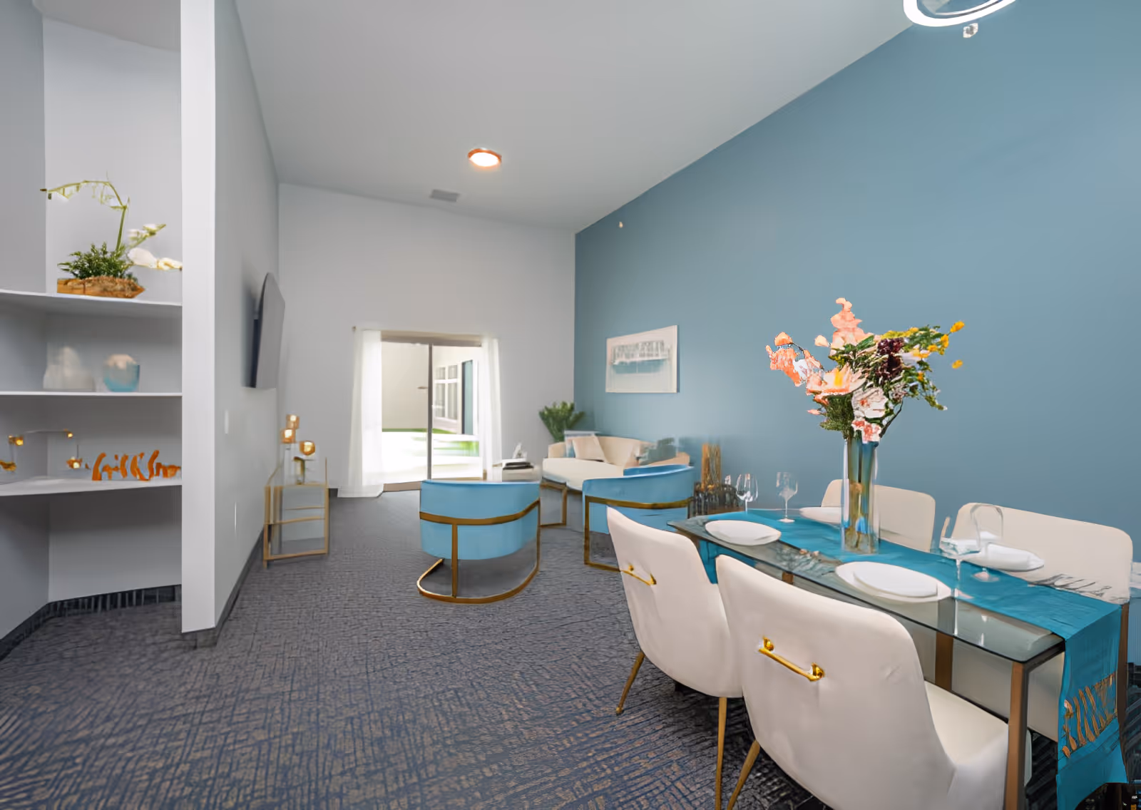 A modern living and dining area with a blue accent wall. The dining table is set with white plates, wine glasses, and a blue table runner, with a vase of flowers as a centerpiece. The living area has light blue chairs with gold accents and a white sofa. There is a sliding glass door with sheer white curtains leading to an outdoor space. Shelves with decorative items are visible on the left side.