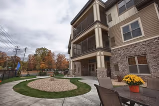 Outdoor patio area of a multi-story assisted living facility with a circular landscaped garden in the center, surrounded by a concrete walkway. There are chairs and a table with a potted plant on the right side, and the building features balconies and large windows. Trees with autumn foliage are visible in the background under a cloudy sky.