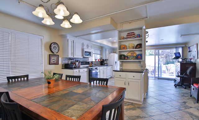 A bright and spacious kitchen and dining area in an assisted living facility. The dining table has a tiled surface with a small potted plant in the center and is surrounded by black chairs. The kitchen features white cabinets, a stove, microwave, refrigerator, and various kitchen appliances. There is a clock and framed picture on the wall near a window with closed blinds. In the background, a sliding glass door leads to an outdoor patio area, and there is a desk with a computer and chair to the right.