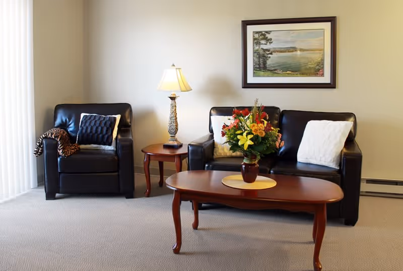 A cozy living room area with a black leather armchair and loveseat, both adorned with white and dark cushions. A wooden coffee table with a floral arrangement in a vase sits in front of the loveseat. A wooden side table with a decorative lamp is positioned between the armchair and loveseat. A framed landscape painting hangs on the beige wall behind the loveseat.