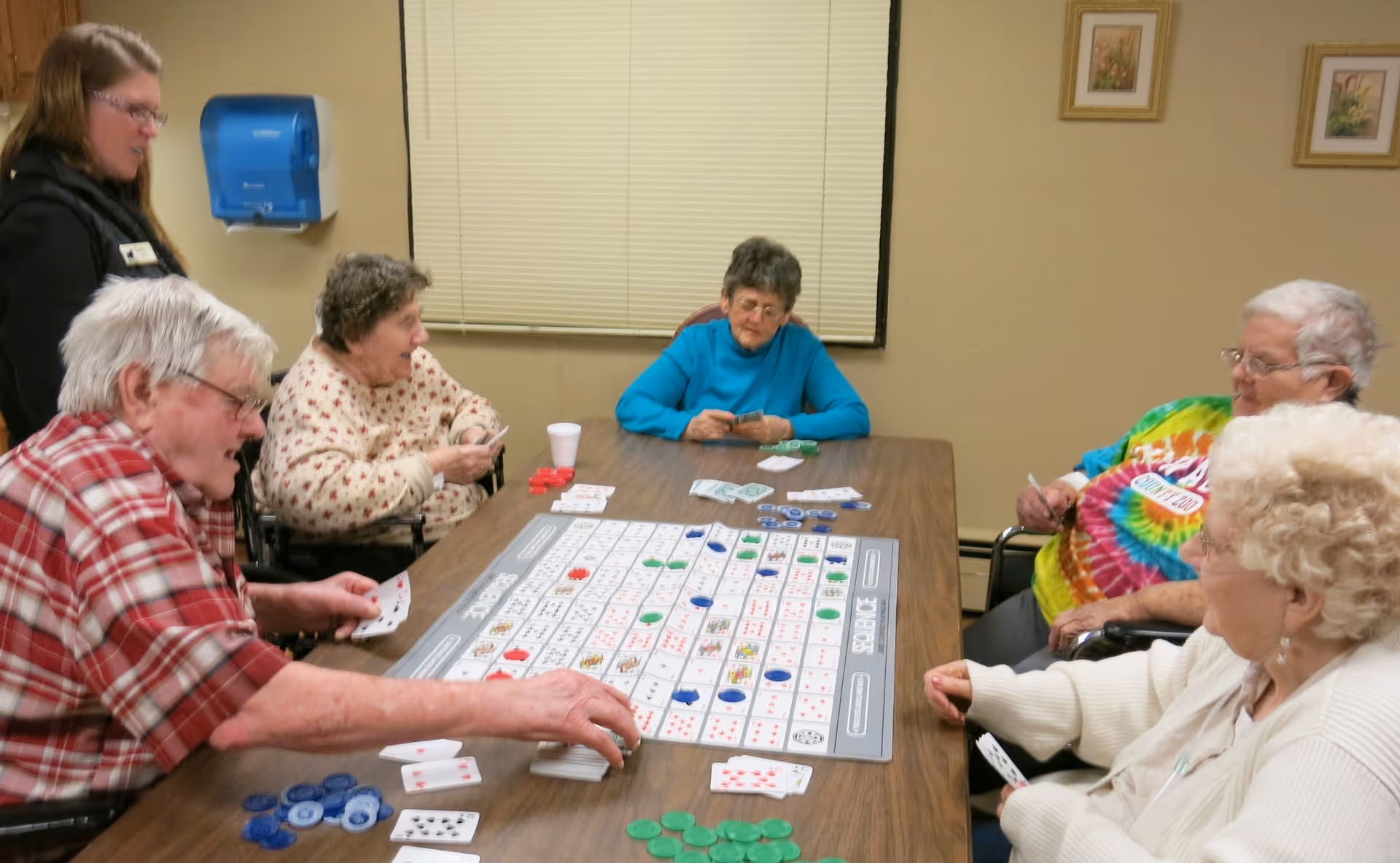 A group of elderly people sitting around a rectangular table playing a board game with cards and colored chips. A staff member stands nearby observing. The room has beige walls with framed pictures and a window with closed blinds.