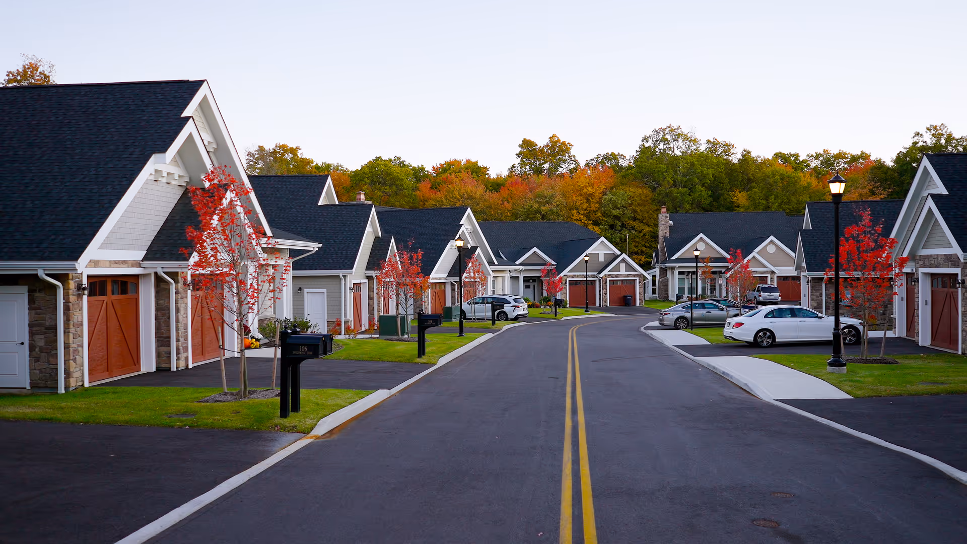 A quiet paved street lined with single-story garage-front cottages, small red-leaved trees, and parked cars in an autumn setting.