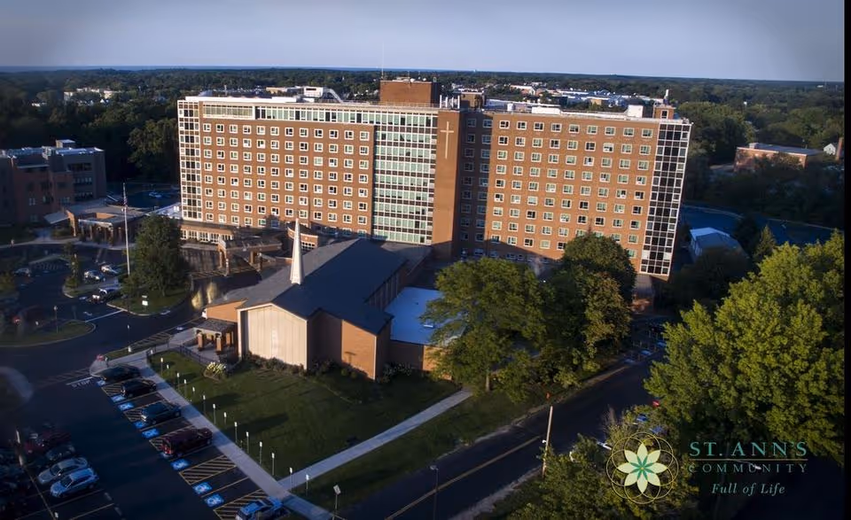 Aerial view of a multi-story senior living building with an attached chapel, parking lot and surrounding trees.