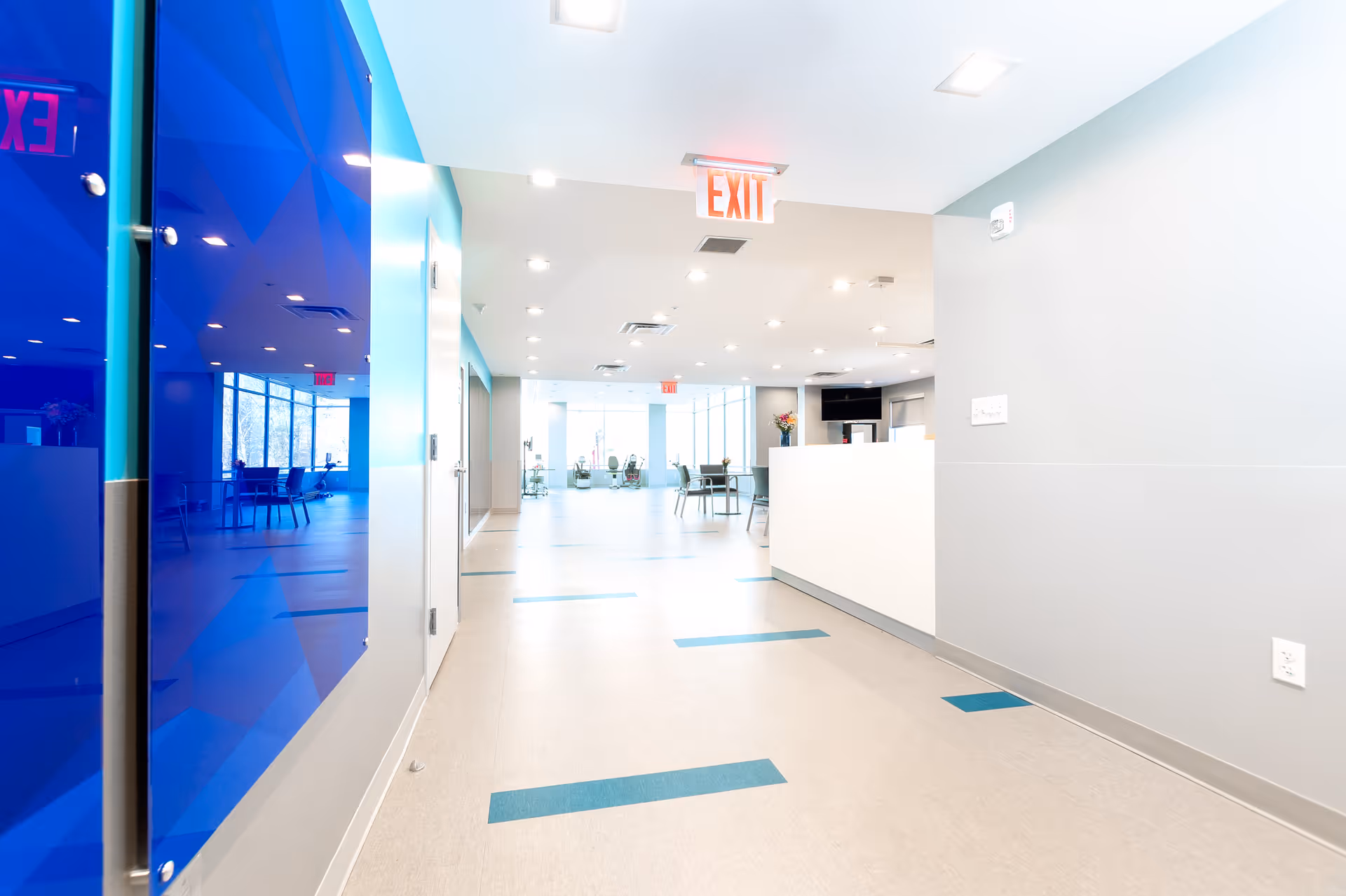 Bright modern hallway and reception area with a white desk, blue glass wall, and seating visible in the distance.