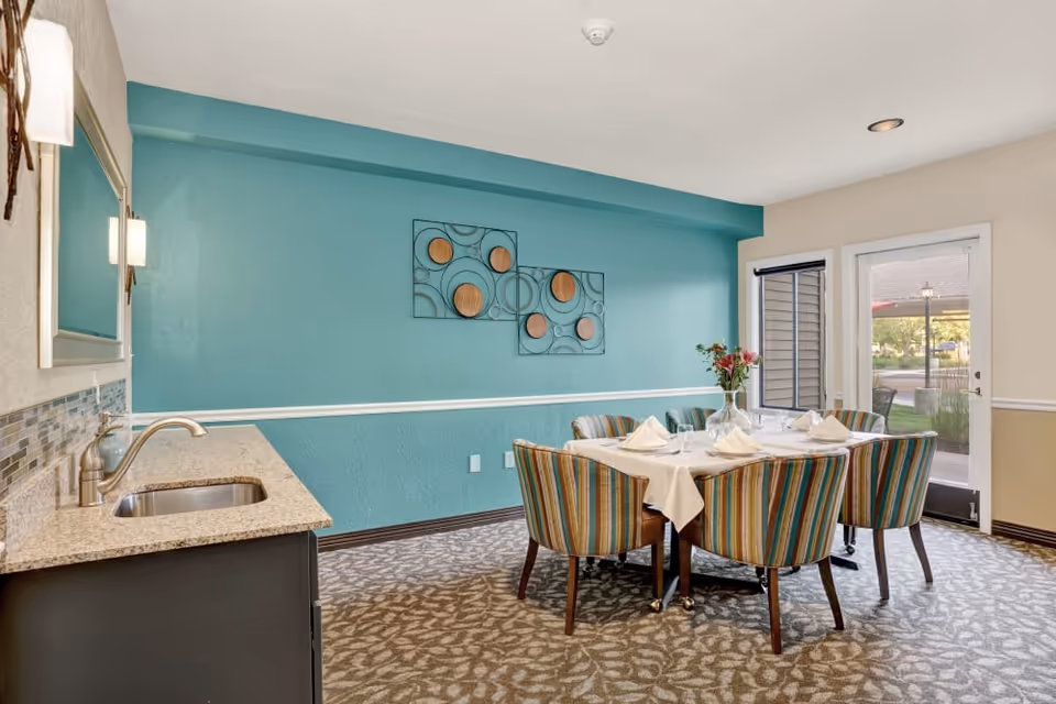 A dining room with a rectangular table set for six people with white napkins and glassware. The chairs around the table have colorful striped upholstery. There is a small countertop with a sink on the left side, and a teal accent wall with decorative wall art. A glass door and window provide a view of the outdoor area with greenery and a patio.