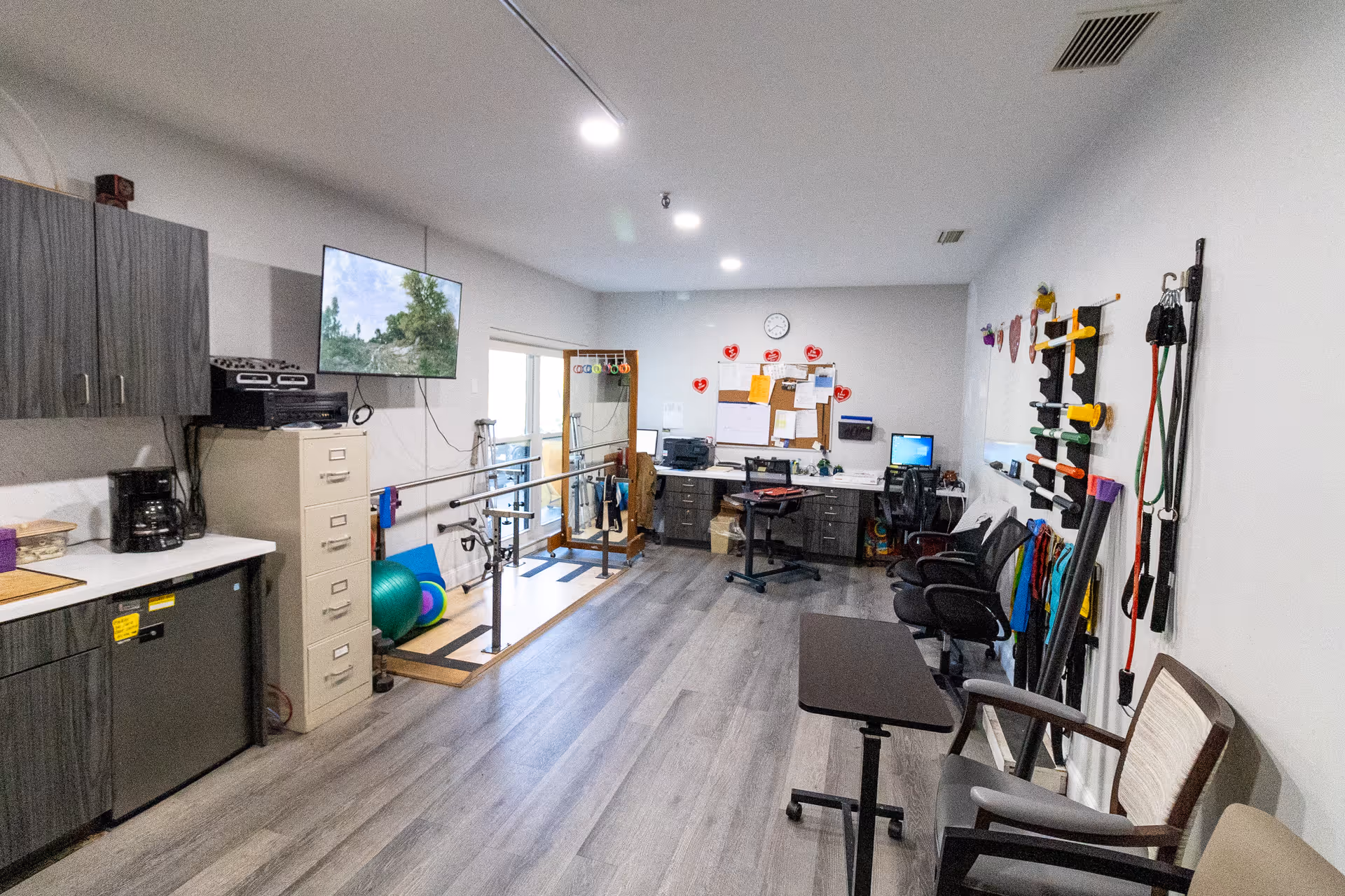 Interior therapy/rehab room with parallel bars, exercise equipment, chairs, desks, and a wall-mounted TV.