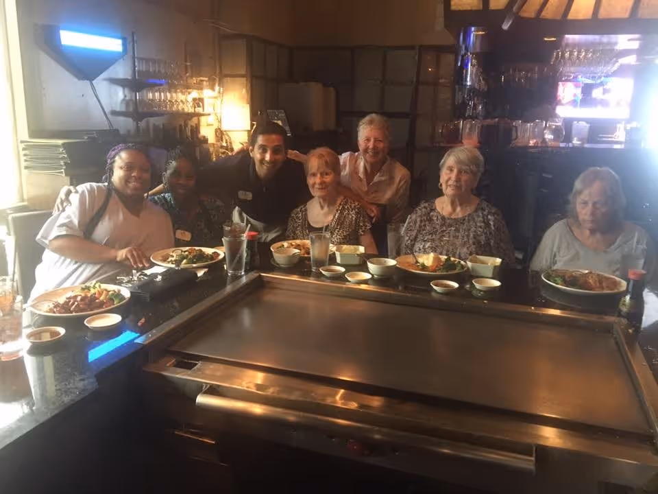 A group of six people, including elderly women and a man, sitting around a teppanyaki grill table in a restaurant with plates of food and drinks in front of them, smiling at the camera.